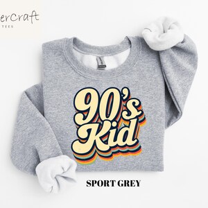 May include: A gray sweatshirt with the text "90's Kid" in a retro rainbow font. The sweatshirt is a crew neck style and is made of a soft, comfortable fabric.