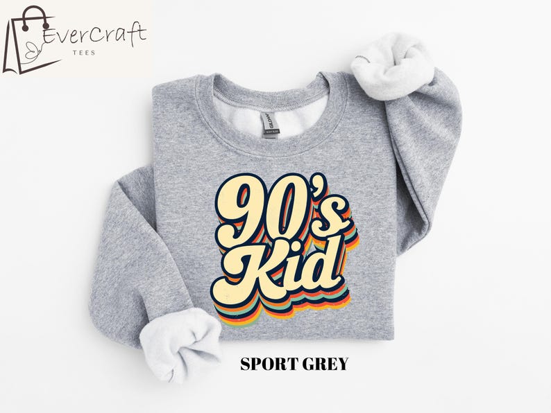 May include: A gray sweatshirt with the text "90's Kid" in a retro rainbow font. The sweatshirt is a crew neck style and is made of a soft, comfortable fabric.