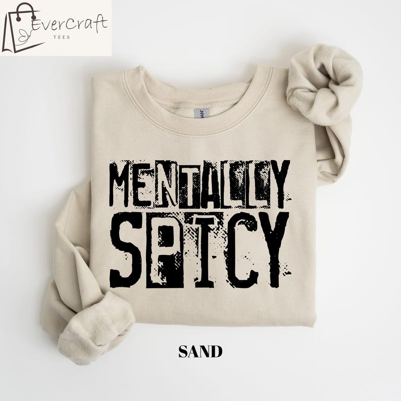 Peut inclure: Un sweat-shirt couleur sable avec les mots "MENTALLY SPICY" imprim&eacute;s en lettres noires et grasses. Le mot "SAND" est imprim&eacute; en dessous. Le sweat-shirt est pli&eacute; proprement sur une surface blanche.