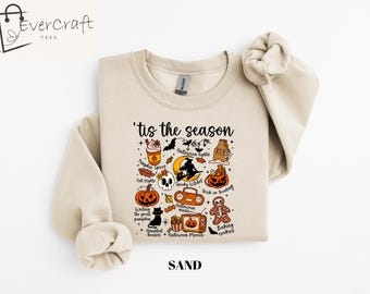 May include: Sand-colored sweatshirt with the text "'tis the season" and Halloween-themed illustrations. The design includes a pumpkin spice latte, bats, a skull, a witch, pumpkins, and a gingerbread man.
