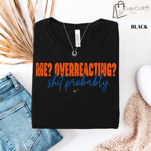 May include: A black t-shirt featuring the orange text "ME? OVERREACTING?" in a distressed block font, followed by the blue cursive text "shit probably." and a small green four-pointed star icon centered below the text on the front of the shirt.