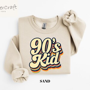 May include: A sand-colored sweatshirt with a retro-style graphic that says "90's Kid" in rainbow colors.