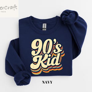 May include: A navy blue sweatshirt with a retro-style graphic that says "90's Kid" in yellow and orange letters. The sweatshirt is a crew neck style.