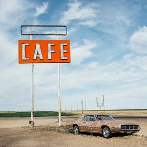 May include: A weathered roadside cafe sign displaying the word "CAFE" in white lettering against an orange backdrop. A rusty classic car is parked on a dirt track beneath a cloudy blue sky.