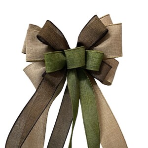 Op de afbeelding: Een grote, decoratieve strik gemaakt van drie verschillende tinten bruin en groen lint. De strik heeft een lange staart en is gebonden in een klassieke, symmetrische stijl.