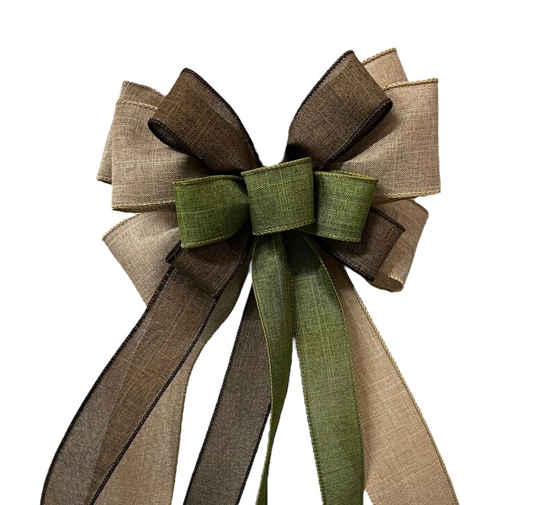 Op de afbeelding: Een grote, decoratieve strik gemaakt van drie verschillende tinten bruin en groen lint. De strik heeft een lange staart en is gebonden in een klassieke, symmetrische stijl.