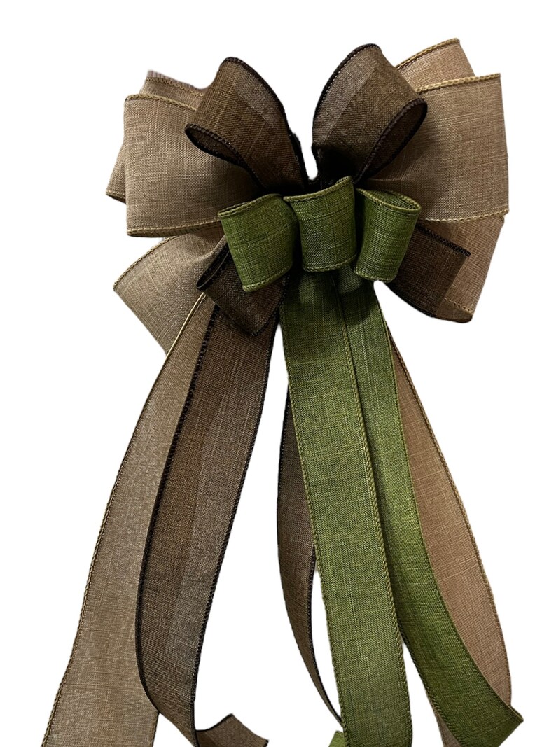 Op de afbeelding: Een grote, decoratieve strik gemaakt van drie verschillende linten. De strik is gemaakt van een bruin en beige lint, een groen lint en een bruin lint met een zwarte rand. De strik is gebonden met een lus en linten.
