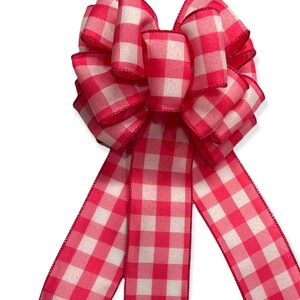May include: A pink and white gingham bow with three tails. The bow is made of a soft, textured fabric and has a classic, country-style design.