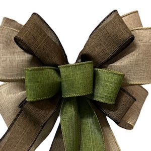 Op de afbeelding: Een grote, meerlagige strik gemaakt van bruin, beige en groen jute lint. De strik is vastgebonden met een bruin lint en heeft een draadlus om op te hangen.