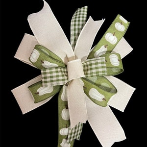May include: A large, decorative bow made from white and green ribbon. The green ribbon has a white pumpkin pattern. The bow is tied in a knot with the tails of the ribbon flowing out from the knot.