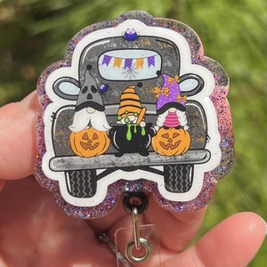 May include: A pink glitter Halloween themed badge reel with a black truck carrying three gnomes. The gnomes are wearing Halloween costumes and holding pumpkins and a cauldron.