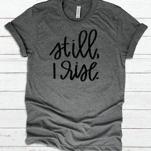 May include: A heather gray t-shirt with the text "still, I rise" printed in black.