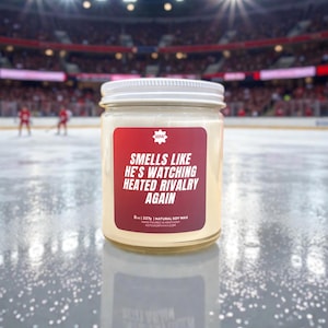 May include: A glass jar candle with a white lid and a red label. The label reads "Smells Like He's Watching Heated Rivalry Again." The candle is on a hockey rink. The candle is 8 oz / 227g.