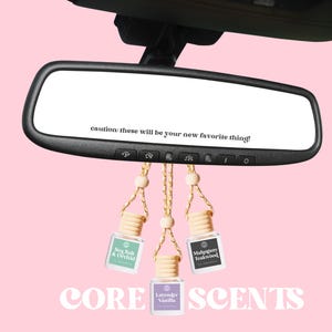 Peut inclure: Trois flacons en verre avec des bouchons en bois et des cordons suspendus à un rétroviseur de voiture. Les flacons sont remplis d'huile parfumée et étiquetés "Sea Salt & Orchid", "Mahogany Teakwood" et "Lavender Vanilla". Le texte "attention : ce seront vos nouveaux préférés !" est visible dans le rétroviseur. Le texte "CORE SCENTS" est imprimé sous les flacons.