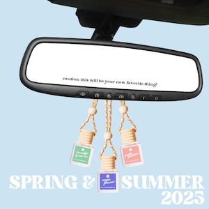 May include: Three car air fresheners hanging from the rearview mirror of a car. The air fresheners are in glass bottles with colorful labels. The labels read "Cucumber Green Tea", "Pink Peony", and "Moon Flower". The text in the rearview mirror reads "caution: this will be your new favorite thing!" The text below the air fresheners reads "SPRING & SUMMER 2025".