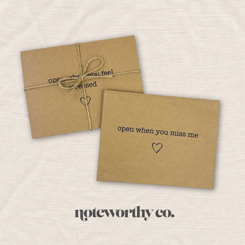 May include: Two brown kraft paper envelopes with handwritten messages. The top envelope says "open when you feel stressed" and the bottom envelope says "open when you miss me". Both envelopes have a small heart drawn on them. The envelopes are tied together with twine. The text "noteworthy co." is printed below the envelopes.