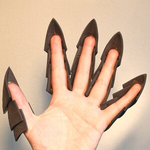 May include: A set of five dark grey foam claw attachments designed to fit over individual fingers. Each piece features a pointed, layered structure that extends beyond the fingertip, creating a sharp, articulated appearance for each digit.