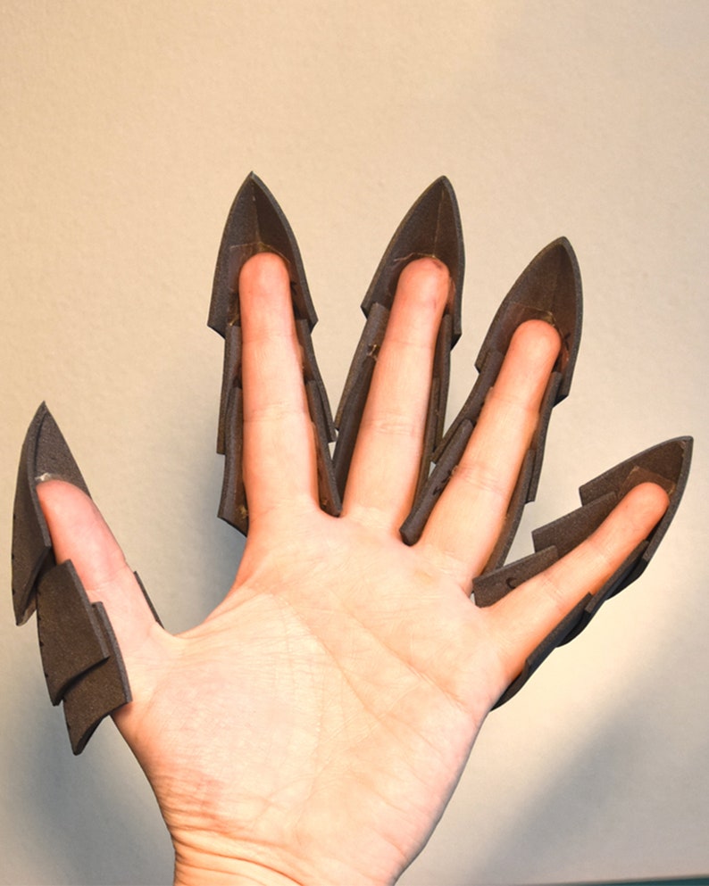 May include: A set of five dark grey foam claw attachments designed to fit over individual fingers. Each piece features a pointed, layered structure that extends beyond the fingertip, creating a sharp, articulated appearance for each digit.