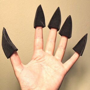 May include: A set of five individual black foam claw attachments designed to fit over the tips of fingers. Each piece is shaped like a pointed, curved talon with a textured surface, providing a costume accessory that covers the fingertip area.