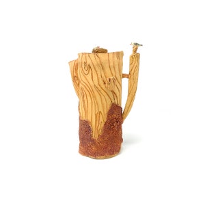 May include: A ceramic pitcher shaped like a tree trunk with a bird perched on a branch. The pitcher is glazed in a light brown colour and has a textured surface.