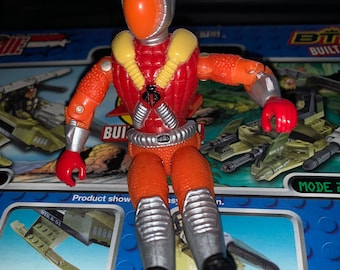 May include: An orange and silver action figure of a soldier wearing a helmet with an orange visor. The figure is holding a gun and has a red and yellow chest piece. The figure is sitting on a toy tank. The background is a blue and green box with the words "Cool Pieces!" and "Launching Missile!" on it.