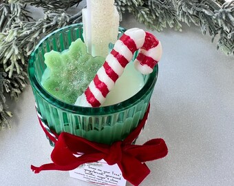 Puede incluir: Un tarro de vidrio verde con una cinta de terciopelo rojo y una etiqueta blanca que dice "Snow me your tree, gingerbread, ground vanilla, sugar & clove". El tarro contiene una vela blanca con un bastón de caramelo rojo y blanco y un copo de nieve verde.