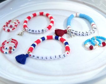 May include: Three beaded bracelets with the word "PHILLIES" spelled out in white letters on black beads. The bracelets are red, white, and blue. There are also two pairs of beaded hoop earrings, one pair is red, white, and blue, and the other pair is blue, white, and red.