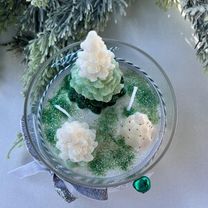 Puede incluir: Una vela en un tarro de vidrio transparente con tres velas con forma de &aacute;rbol de pino verde y blanco. El tarro est&aacute; lleno de purpurina verde.