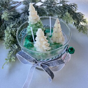 Puede incluir: Una vela de vidrio en forma de tarro con tres velas con forma de &aacute;rbol de Navidad blancas y verdes en su interior. El tarro est&aacute; decorado con una cinta plateada y blanca.