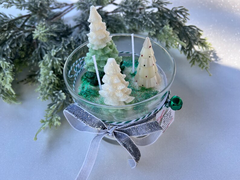 Puede incluir: Una vela de vidrio en forma de tarro con tres velas con forma de &aacute;rbol de Navidad blancas y verdes en su interior. El tarro est&aacute; decorado con una cinta plateada y blanca.