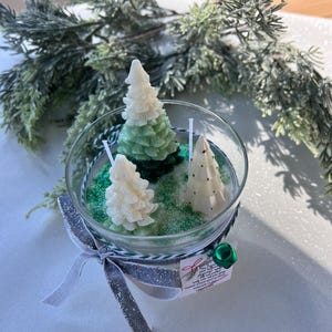 Puede incluir: Una vela en un tarro de vidrio transparente con tres velas de &aacute;rbol de Navidad verde y blanco en su interior. El tarro est&aacute; lleno de purpurina verde y los &aacute;rboles est&aacute;n rodeados de purpurina blanca. El tarro est&aacute; atado con una cinta plateada y blanca y una campana verde.