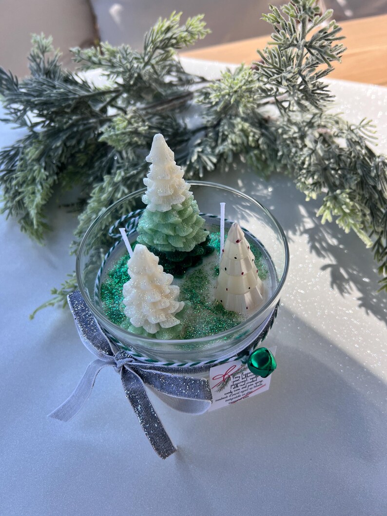 Puede incluir: Una vela en un tarro de vidrio transparente con tres velas de &aacute;rbol de Navidad verde y blanco en su interior. El tarro est&aacute; lleno de purpurina verde y los &aacute;rboles est&aacute;n rodeados de purpurina blanca. El tarro est&aacute; atado con una cinta plateada y blanca y una campana verde.