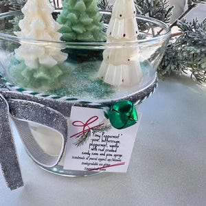 Puede incluir: Una vela en un tarro de vidrio con una cinta plateada y verde y un amuleto de campana verde. La vela est&aacute; decorada con tres velas con forma de &aacute;rbol de pino blanco y verde. El tarro tiene una etiqueta que dice "Piney Peppermint - pino, crema de mantequilla, menta, vainilla con ca&ntilde;a de az&uacute;car triturada real y ramitas de pino. Hecho a mano y vertido con soja org&aacute;nica/cera de abejas. Brillo ecol&oacute;gico biodegradable. Esencias esenciales."
