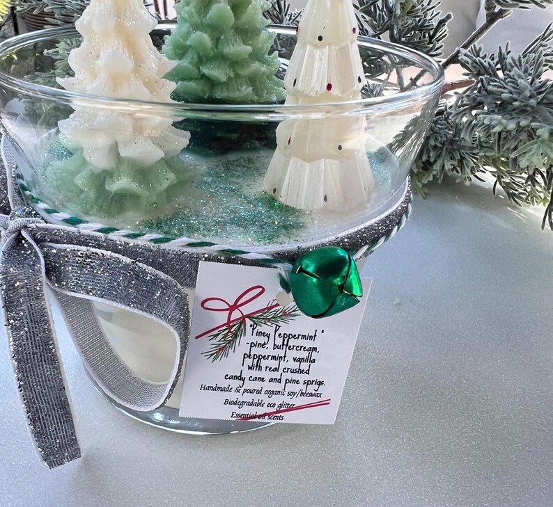 Puede incluir: Una vela en un tarro de vidrio con una cinta plateada y verde y un amuleto de campana verde. La vela est&aacute; decorada con tres velas con forma de &aacute;rbol de pino blanco y verde. El tarro tiene una etiqueta que dice "Piney Peppermint - pino, crema de mantequilla, menta, vainilla con ca&ntilde;a de az&uacute;car triturada real y ramitas de pino. Hecho a mano y vertido con soja org&aacute;nica/cera de abejas. Brillo ecol&oacute;gico biodegradable. Esencias esenciales."