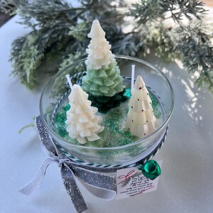 Puede incluir: Una vela de vidrio en forma de tarro con tres velas de &aacute;rbol de Navidad blancas y verdes en su interior. El tarro est&aacute; decorado con purpurina verde y una cinta plateada y blanca con un dije de campana verde. La etiqueta dice "Tiny Evergreen" con un mensaje escrito a mano sobre la vela.