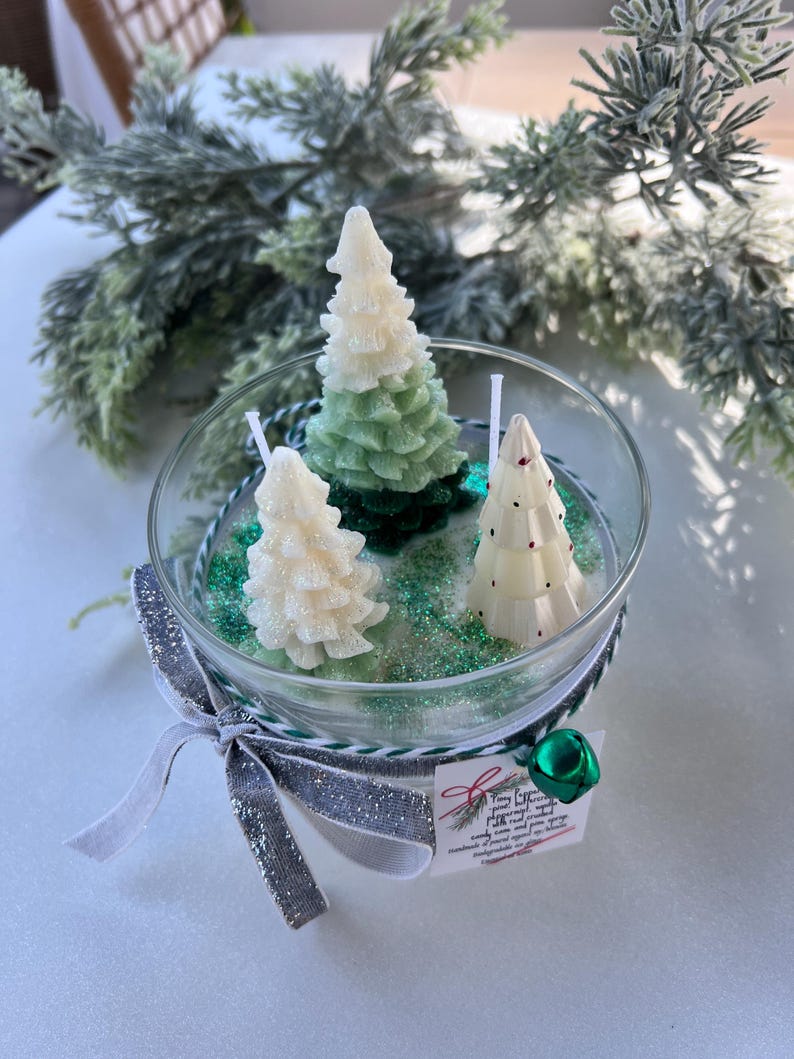 Puede incluir: Una vela de vidrio en forma de tarro con tres velas de &aacute;rbol de Navidad blancas y verdes en su interior. El tarro est&aacute; decorado con purpurina verde y una cinta plateada y blanca con un dije de campana verde. La etiqueta dice "Tiny Evergreen" con un mensaje escrito a mano sobre la vela.