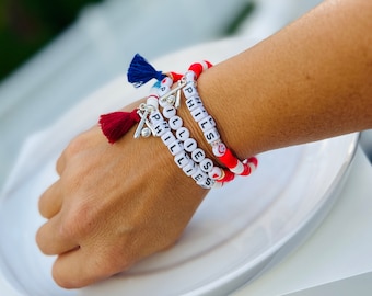 May include: Three white beaded bracelets with the words "I Love Phillies" and "Phillies" on them. The bracelets are stacked on a person's wrist and have red and white beads with a small silver baseball bat charm.
