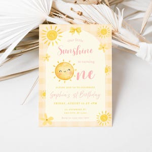 May include: A rectangular invitation card with a yellow and white gingham pattern. The card features a smiling sun illustration and the text "Our little Sunshine is turning one." Additional text includes the birthday details.