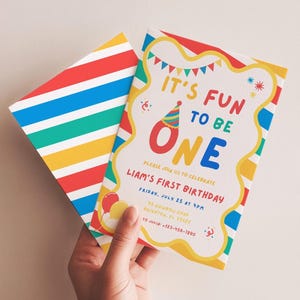Op de afbeelding: Een uitnodigingskaart voor een eerste verjaardag met bijpassende envelop, voorzien van horizontale regenboogstrepen in rood, blauw, groen en geel. De kaart bevat de tekst IT'S FUN TO BE ONE, LIAM'S FIRST BIRTHDAY en feestdetails met kleurrijke ballonnen.