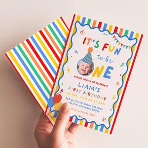 May include: A first birthday invitation card featuring a photo of a baby in a party hat, colorful text reading IT'S FUN TO BE ONE, LIAM'S FIRST BIRTHDAY, and party details. The card has a wavy blue border and a rainbow striped envelope back.