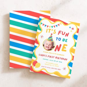 May include: A birthday invitation card featuring a colorful diagonal striped pattern in red, yellow, blue, and teal. The card displays the text IT'S FUN TO BE ONE, a photo of a baby wearing a party hat, and event details for Liam's first birthday.