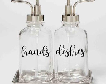 May include: Two clear glass soap dispensers with silver pumps and labels that say "hands" and "dishes". The dispensers are sitting on a silver tray.