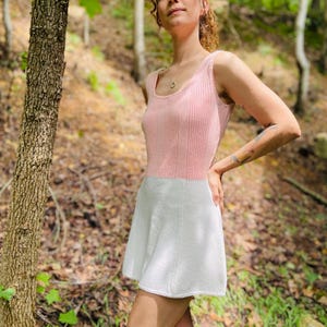 May include: A woman wearing a pink and white knitted dress in a forest setting. The dress has a sleeveless bodice and a flared skirt.