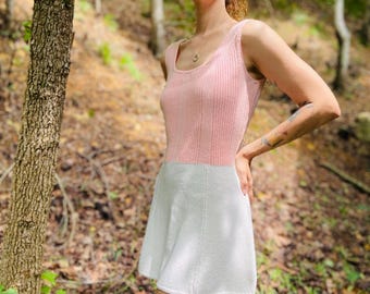 May include: A woman wearing a pink and white knitted dress in a forest setting. The dress has a sleeveless bodice and a flared skirt.