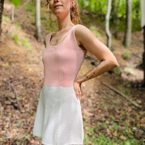 May include: A pink and white knitted dress with a sleeveless bodice and a flared skirt. The dress is worn by a person standing in a forest setting.