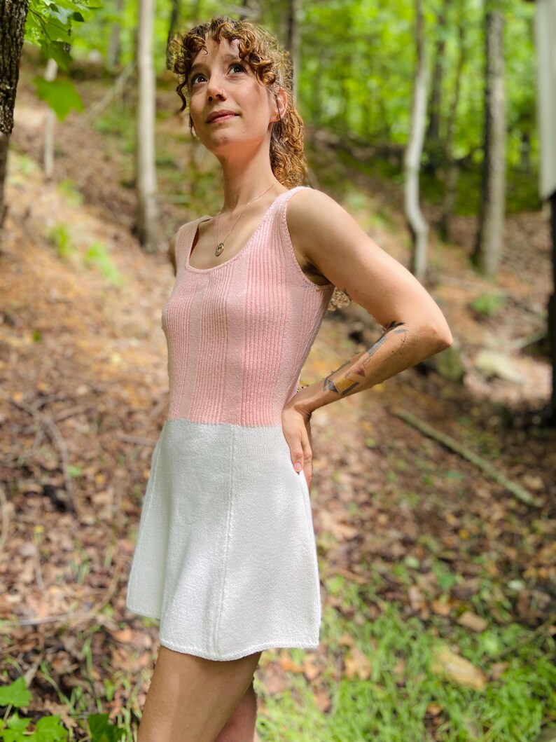 May include: A pink and white knitted dress with a sleeveless bodice and a flared skirt. The dress is worn by a person standing in a forest setting.