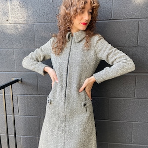 May include: A vintage-style dress in a black and white herringbone pattern. The dress features long sleeves, a collared neckline, and a full-length zipper. Two small pockets are visible on the front. The dress is knee-length.