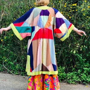 May include: A colourful patchwork kimono jacket with a variety of fabric pieces in shades of red, blue, green, and tan. The jacket has wide sleeves and a yellow trim. The person is wearing a patterned skirt and purple shoes.
