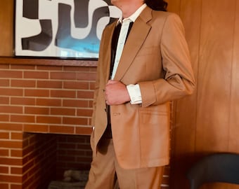 May include: A man wearing a tan suit with a white shirt and a black tie. He is standing in front of a brick fireplace with a black and white abstract painting above it.