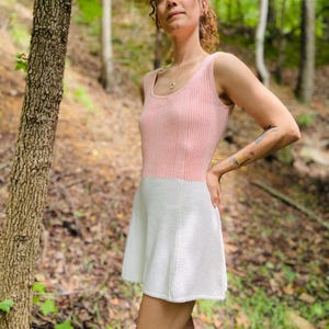 May include: A pink and white knitted dress with a sleeveless bodice and a flared skirt. The dress is worn by a person standing in a forest.